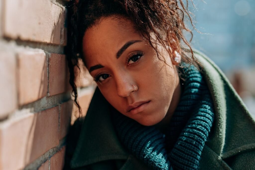 A thoughtful woman in a green coat leans against a brick wall on a sunny day.